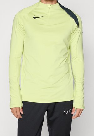 Homme portant un haut Nike à manches longues vert clair avec col zippé et un pantalon Nike noir, debout devant un fond uni.