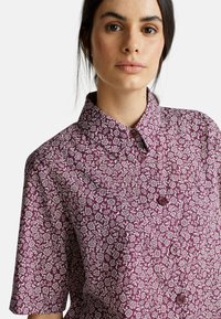 Donna con capelli scuri che indossa una camicia a maniche corte con bottoni, con un motivo floreale viola e bianco, su uno sfondo neutro.