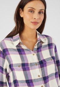Camisa de cuadros morados y blancos con cuello, cierre de botones en la parte delantera y dos bolsillos en el pecho. Fabricada con una tela suave y texturizada.