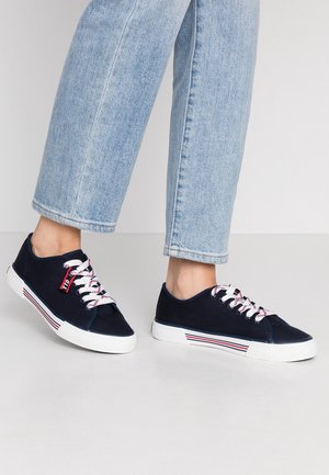 Osoba ubrana w jasnoniebieskie dżinsy oraz granatowe niskie trampki z białą podeszwą i sznurowadłami z czerwonymi i czarnymi akcentami.