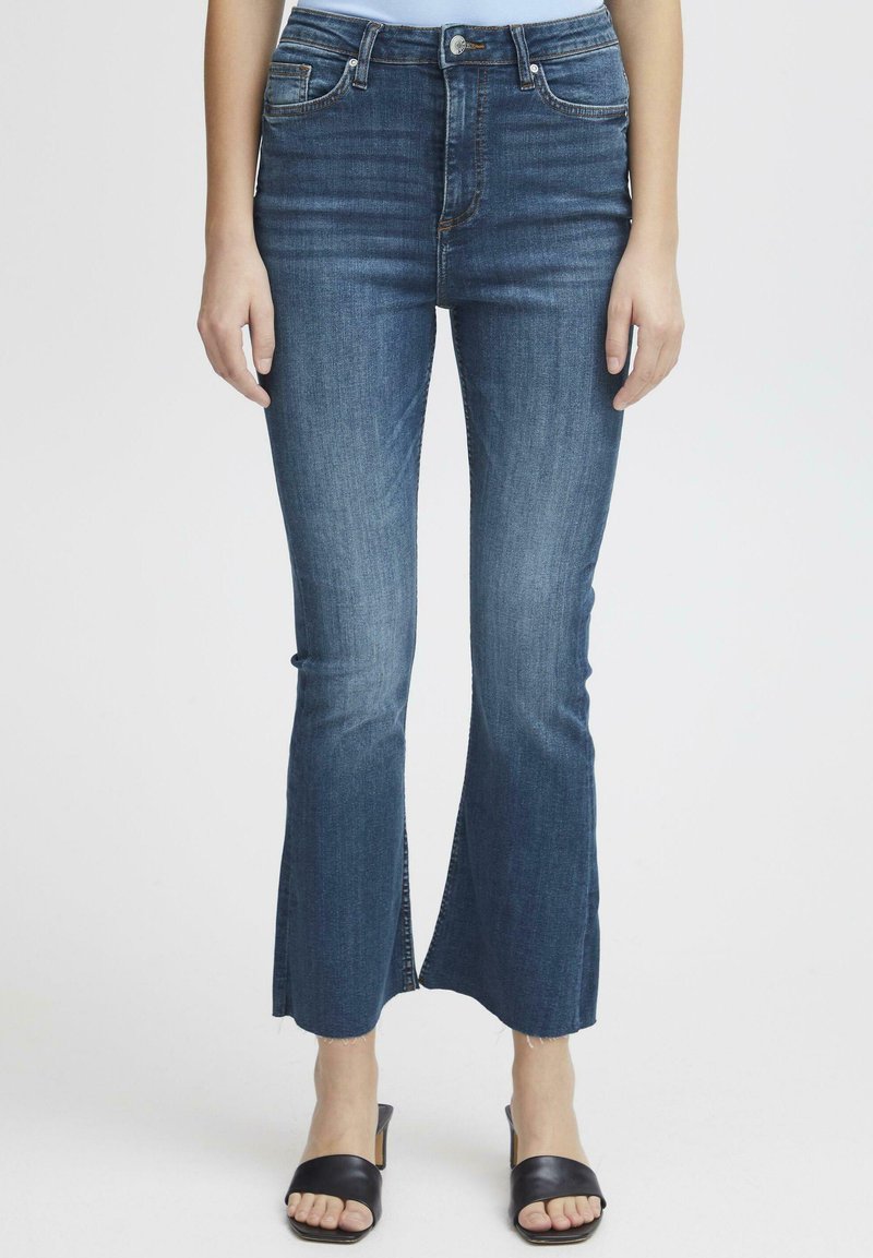 Jeans in denim a svasatura ampia di blu medio con vita alta, caratterizzati da fessure laterali all'orlo e un leggero effetto scolorito. Abbinati a sandali neri a ciabatta.