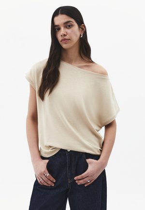 Femme aux longs cheveux foncés portant un haut beige ample tombant sur les épaules et un jean foncé, les mains dans les poches, debout devant un fond blanc uni.