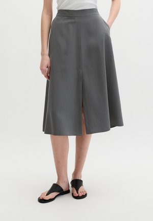 Woman wearing a gray knee-length A-line skirt with front slit, white top, and black open-toe sandals, standing against a plain background.