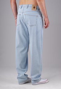 Man wearing light blue loose-fit jeans and white sneakers standing with back facing camera against plain background.