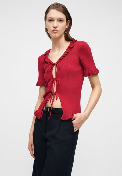 Mujer con un top rojo de punto de manga corta con volantes y lazos delanteros, combinado con pantalones negros y pendientes de aro dorados.