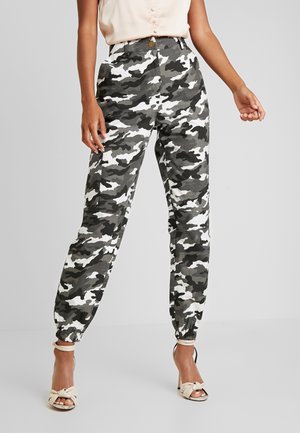 Kobieta w czarno-biało-szarych, moro, wysokich spodniach cargo z elastycznymi mankietami oraz beżowych sandałach na obcasie z odkrytymi palcami i paskiem wokół kostki.