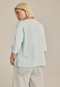 Blonde Person, der ein lockeres hellblaues T-Shirt und hellgraue Jeans trägt, steht leicht gedreht und schaut zur Seite.