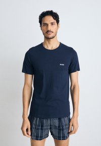 T-shirt de algodão azul-marinho com decote redondo e mangas curtas, apresentando um pequeno logo branco "BOSS" no peito. Usado com calções xadrez.