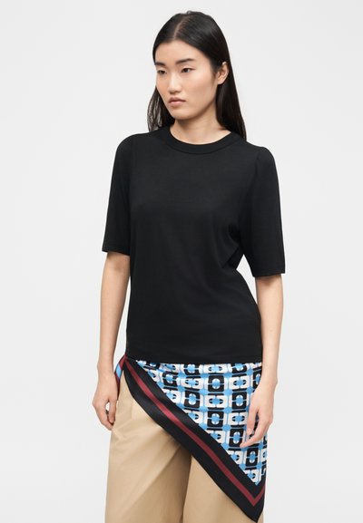 Femme portant un haut noir à manches courtes et un pantalon beige, avec une écharpe à motifs géométriques bleus, noirs et rouges nouée autour de la taille.