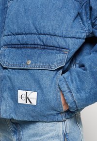 Jasnoniebieska kurtka dżinsowa z pikowanym wzorem, wyposażona w kieszeń na zatrzask oraz białą metkę z logo "Calvin Klein Jeans".
