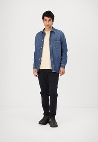 Blauwe denim shirt met lange mouwen, knoopsluiting aan de voorkant, enkele borstzak, gedragen over een lichtbeige T-shirt en zwarte broek, gecombineerd met donkere sneakers.