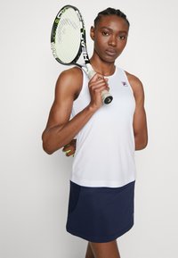 Hvit ermeløs tennis topp med en logo, kombinert med en marineblå skjørt. Holder en tenniskølle med teksturert grep og lysegrønne detaljer.