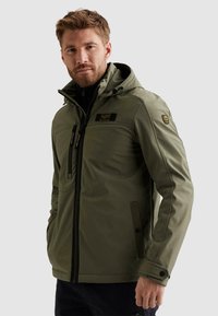 Uomo con giacca con cappuccio verde oliva con cerniere nere e toppe, in piedi davanti a uno sfondo grigio chiaro uniforme.