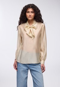 Blusa di seta crema con colletto a cravatta, maniche lunghe e forma fluida. Il tessuto è liscio e semitrasparente. Abbinata a jeans azzurri chiari.
