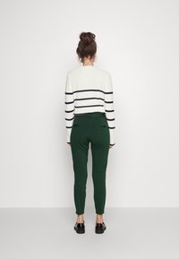 Biało-granatowy prążkowany krótki sweter w połączeniu z zielonymi dopasowanymi spodniami, model tyłem, płaskie czarne buty, gładka tekstura materiału.