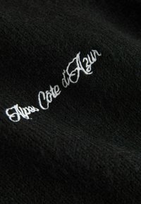 Zwarte stof met een textuur en witte geborduurde tekst 'Alpes Côte d'Azur'.