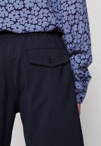 Chemise bleue fleurie à manches longues glissée dans un pantalon bleu marine avec une poche arrière boutonnée, montrant le bas du dos et le bras du porteur.