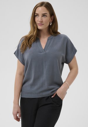 Femme aux cheveux châtains clairs portant des boucles d'oreilles argentées en anneau, un haut gris à manches courtes et col en V ample, et un pantalon noir, debout avec une main dans la poche.