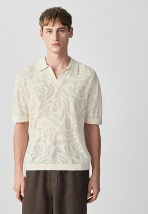 Junger Mann, der ein cremefarbenes kurzärmeliges Strick-Poloshirt mit floralem Muster und braune Hosen trägt, steht vor einem schlichten hellen Hintergrund.