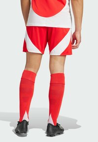 Rode sportieve shorts met witte gebogen accenten, gecombineerd met kniehoge rode sokken en zwarte voetbalschoenen. Het gaasstof biedt ademend vermogen.