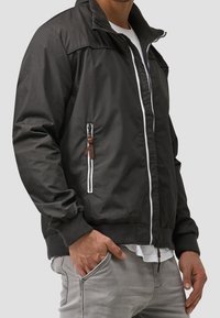 Veste bomber noire avec fermeture éclair à l'avant, poignets côtelés et poches. Elle est dotée de fermetures éclair blanches et d'un accent en cuir marron sur la poche latérale.
