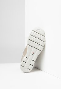 Zapatilla con parte superior beige, diseño texturizado y suela de goma blanca que presenta patrones de relieve distintivos y un acento de logo.