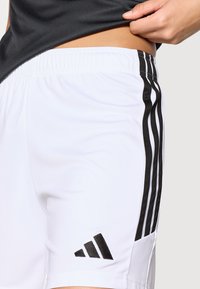 Λευκά αθλητικά σορτς με μαύρες ρίγες στα πλευρά, ελαστική μέση και το λογότυπο της Adidas σε μαύρο κοντά στο τελείωμα. Λείο, ελαφρύ ύφασμα.