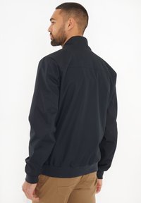 Giacca nera con zip e colletto alto, polsini e vita a costine; presenta una texture liscia e una vestibilità comoda, indossata sopra pantaloni beige.
