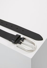 Schwarzer Leder-Gurt mit einer silbernen Metallschnalle, der mehrere Einstelllöcher aufweist und eine glatte, flache Textur hat.
