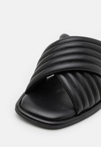 Sandale à glissière noire avec des brides rembourrées cousues horizontalement et une semelle carrée légèrement rembourrée.