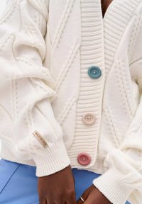 White knitted cardigan featuring ribbed texture, three buttons in blue, pink, and beige, with a labeled tag on the sleeve.