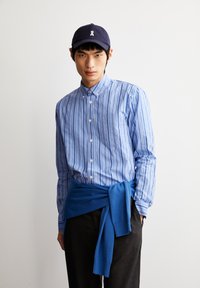 Blauw gestreepte button-up overhemd, marineblauwe pet met logo, blauwe trui om de taille geknoopt, zwarte broek, gladde stof, casual ontwerp.