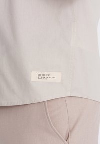 Hellgraues Hemd aus glattem Stoff mit geradem Saum, das am unteren Rand ein gesticktes Etikett mit der Aufschrift "OMBRA STREETSTYLE seit 1985" trägt.