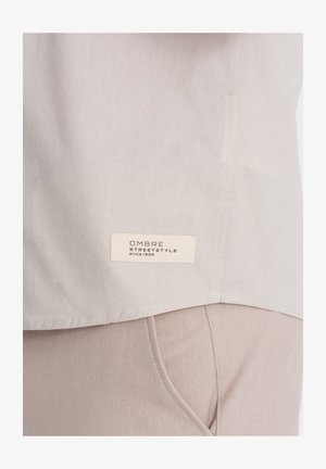 Jasnoszara koszula z gładkiego materiału i prostym dołem, z haftowaną metką oznaczającą "OMBRA STREETSTYLE since 1985" na dolnej krawędzi.