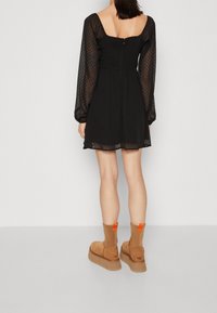 Svart klänning med genomskinliga langärmade prickar, fyrkantig halsringning och figurnära liv. Bärs med tanfärgade ankelstövlar med en orange accent.