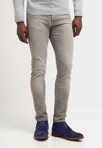 Jeans slim-fit grigio chiaro in denim, abbinati a stivaletti alla caviglia in suede blu con lacci blu. Il pullover è grigio chiaro, con scollatura a girocollo.