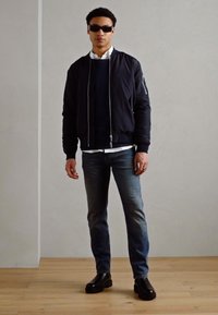 Black bomber jacket, navy sweater, white shirt, blue jeans, and black shoes. Model wears sunglasses, standing against a plain background.