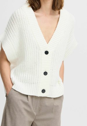 Femme portant un cardigan sans manches blanc en tricot avec des boutons noirs et un pantalon beige à fines rayures, les mains dans les poches.
