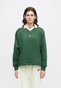 Pessoa a usar um sweatshirt verde com o logótipo "NIKE" e calças creme com o swoosh da Nike, em pé contra um fundo branco liso.
