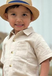 Ljusbeige kortärmad skjorta med krage, en bröstficka och fyra gyllene knappar. Bärs med en halmhatt med ett blått band.