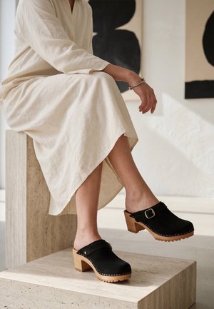 Személy krémszínű ruhát és ezüst karkötőt visel, egy kőblokkra ülve, fekete fa talpú klumpákat csatolva mutat be.