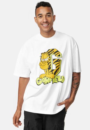 T-shirt oversize blanc avec un graphisme de Garfield, un "G" rayé jaune et noir et le texte "GARFIELD" en vert en bas.