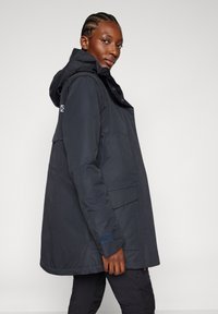 Kvinna klädd i en mörk huva vinterkappa med fickor, tittar över sin axel mot en enkel ljus bakgrund.