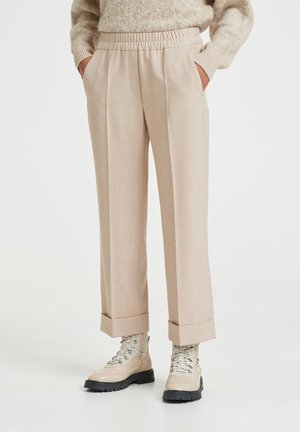 Lys beige, skræddersyede bukser med plisseret design, elastisk talje og opsmøgede ender, parret med beige snørebåndsstøvler på en hvid baggrund.