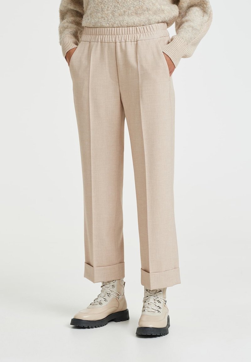 Vaaleanbeige mukautetut housut, joissa on laskostettu muotoilu, joustavyö, ja käänteet, yhdessä vaaleanbeigejen nauhakuormanhousujen kanssa valkoista taustaa vasten.