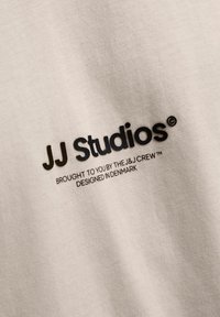 Tela de algodón beige con una textura suave que presenta texto impreso en negro: "JJ Studios" y texto adicional debajo, incluyendo "Te lo trae..."