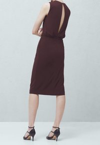 Brązowa sukienka do kolan z wycięciem na plecach, bez rękawów i elastycznym pasem. Noszona z czarnymi szpilkami na kostce.