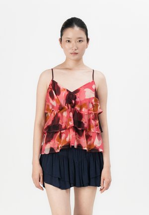 Femme portant un top floral sans manches, à volants, rose et rouge, avec des bretelles fines, et une jupe bleue marine à plusieurs couches, se tenant devant un fond uni.