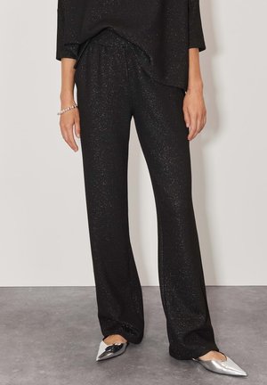 Pantalon large noir pailleté porté avec un haut assorti et des ballerines argentées à bouts pointus sur un mannequin devant un fond uni.