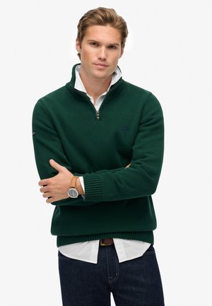 Suéter de punto verde oscuro con cuello con cremallera, puños y dobladillo acanalados; llevado sobre una camisa blanca; el modelo está de pie con los brazos cruzados.